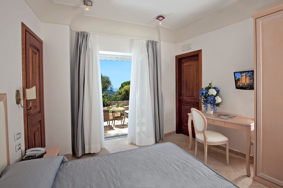 A hotel room with a bed, a desk with a chair, a TV, and a balcony with a view.