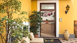 A hotel entrance with a yellow wall, a large potted plant, a beige sofa, and a welcome mat. - Villa Ceselle (Anacapri)