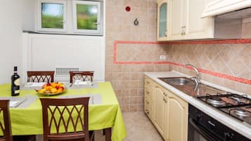 Une cuisine avec une table à manger, des chaises, un panier de fruits, une bouteille de vin et une cuisinière.