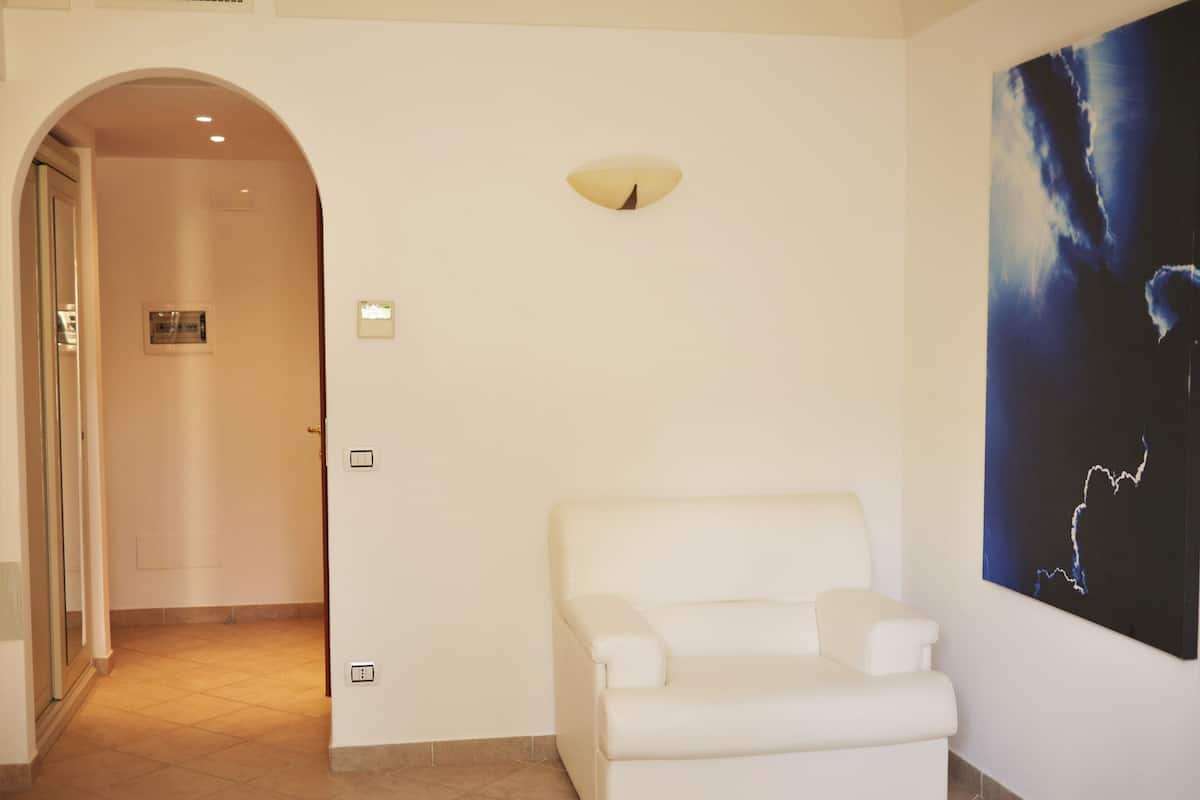 A room with a white armchair, a door with an intercom, a light switch, and a wall-mounted light fixture.