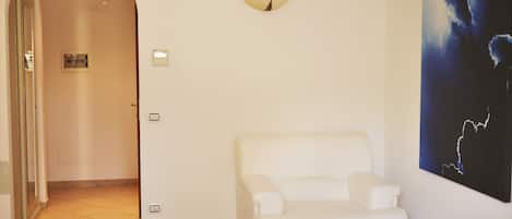 Una habitación con un sillón blanco, una puerta con interfono, un interruptor de luz y una lámpara de pared.