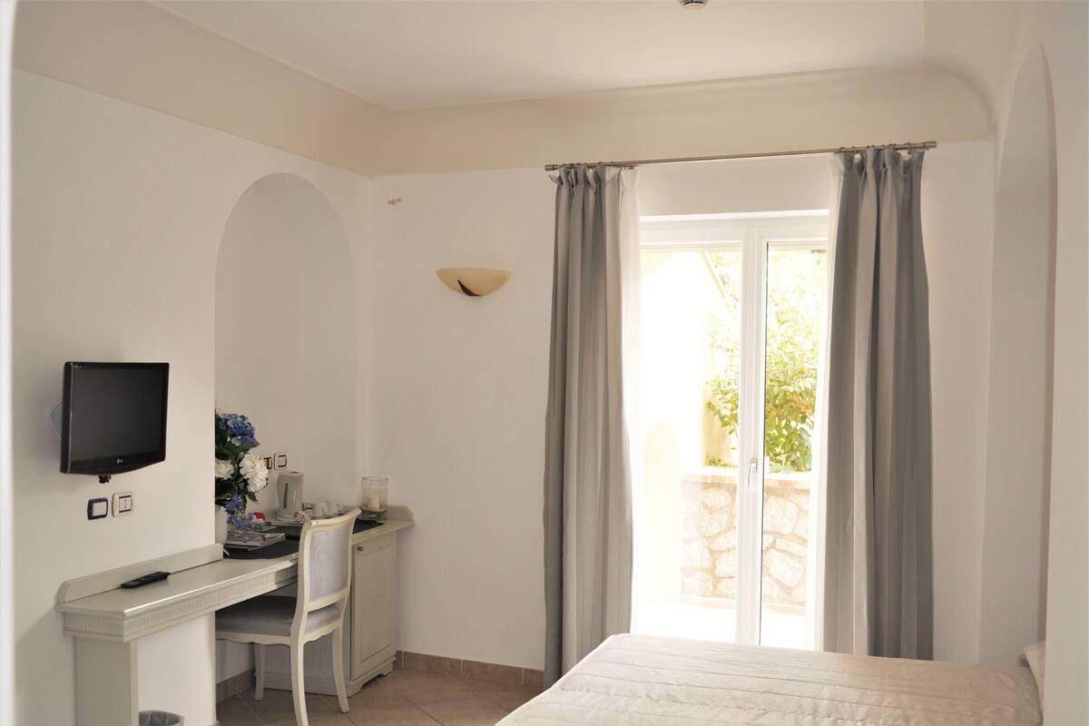 A bedroom with a bed, a desk with a chair, a TV, and a window with curtains.