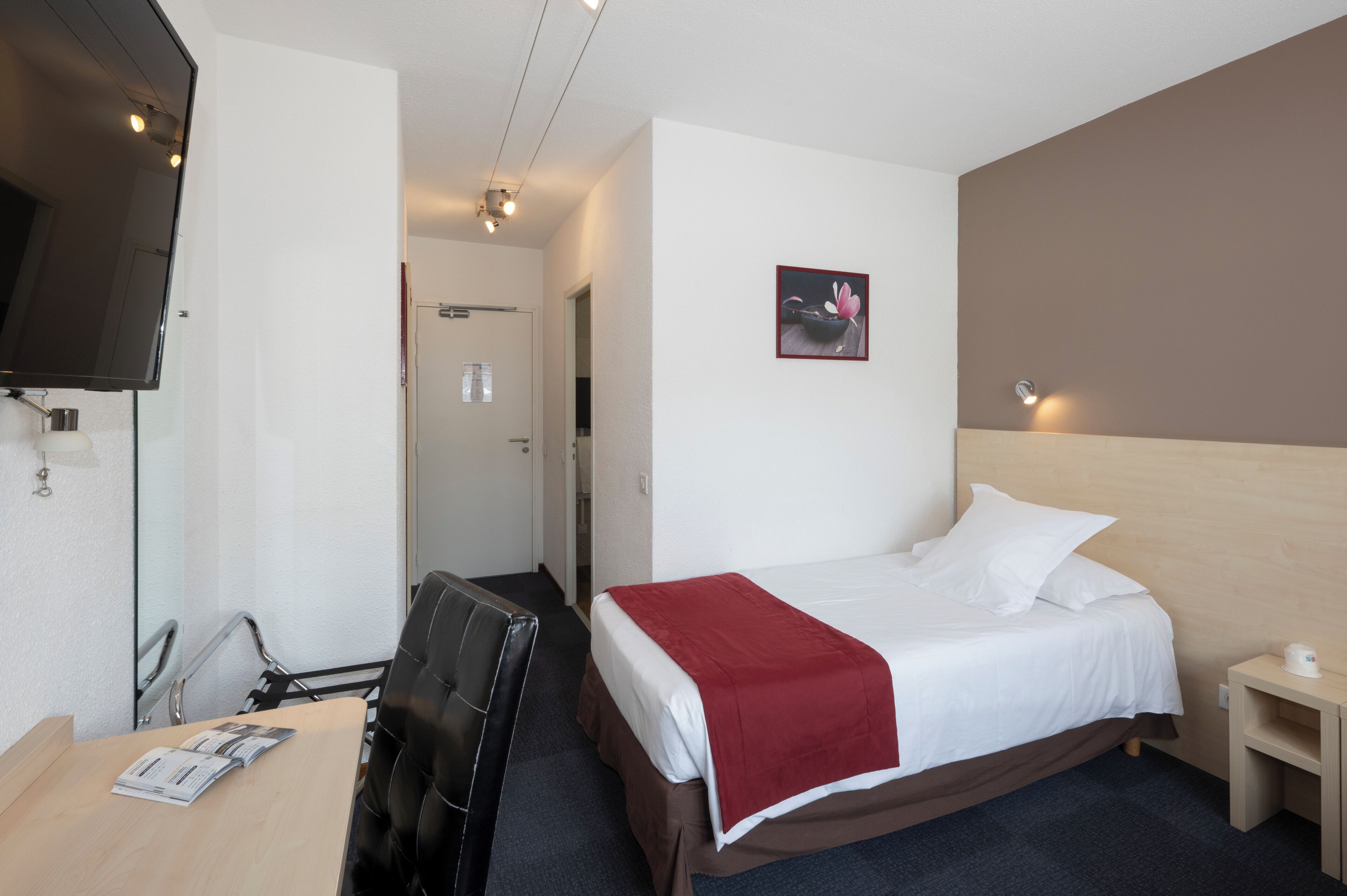 Quarto de hotel com cama, escrivaninha com cadeira, televisão fixada na parede e mesa de cabeceira pequena.