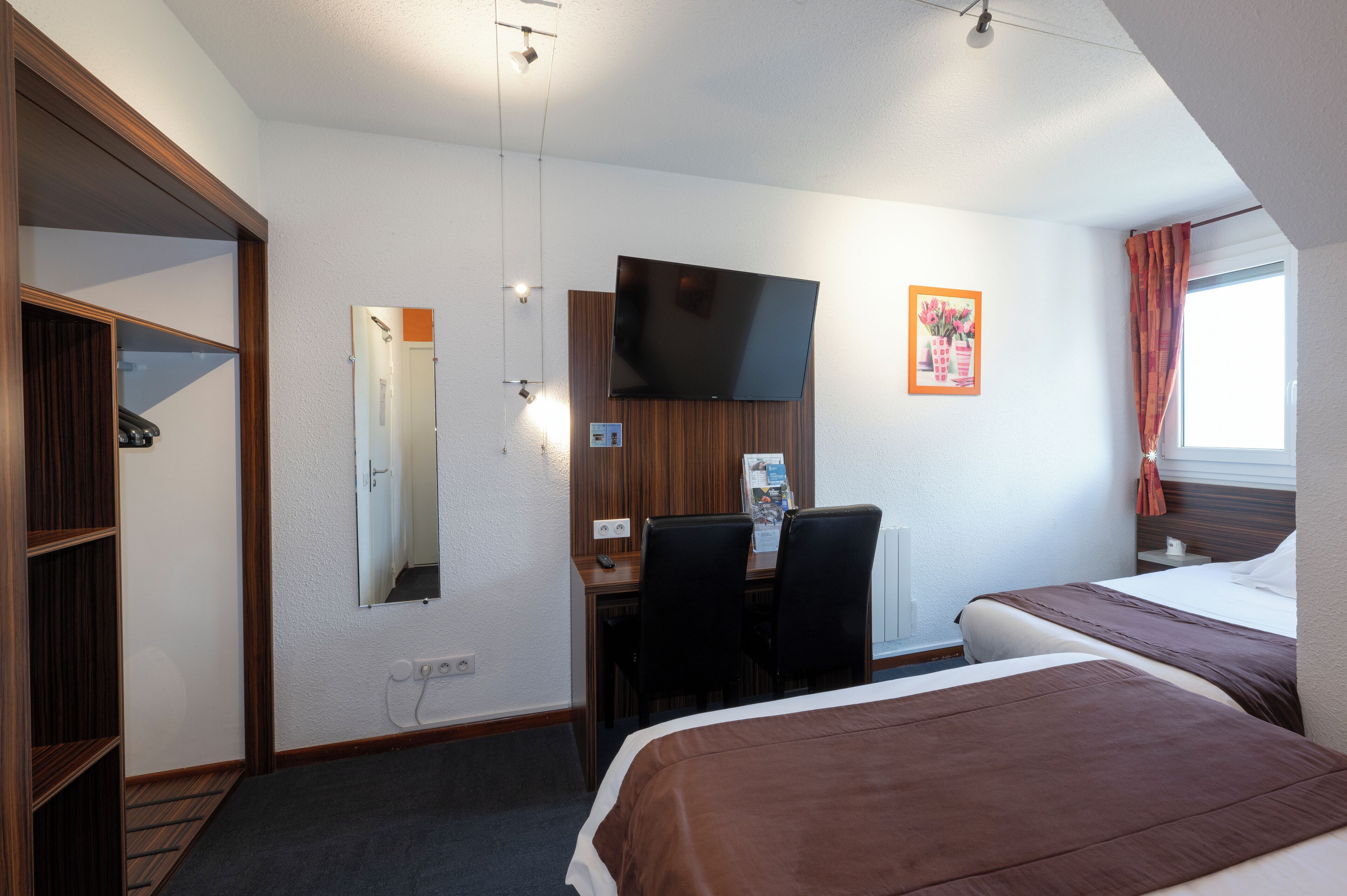 A hotel room with a bed, a desk with a chair, a television mounted on the wall, and a window with curtains.