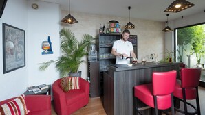 Uma área de bar com um bartender, cadeiras vermelhas, uma planta em vaso e uma estante com garrafas.