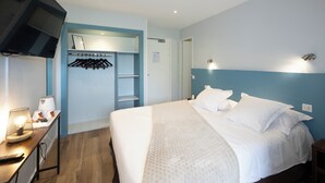 Quarto de hotel com cama, televisão na parede, secretária com luminária e cabideiro com ganchos.