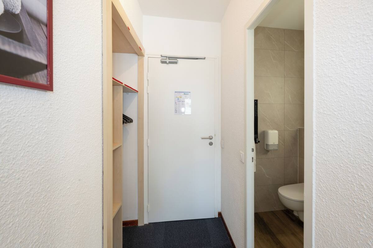 Un pasillo estrecho con una puerta blanca, un estante pequeño con un objeto rojo y un baño con un inodoro y dispensador de toallas de papel.