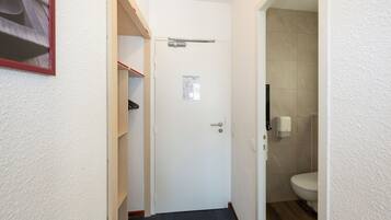 A narrow hallway with a white door, a small shelf with a red object, and a bathroom with a toilet and a paper towel dispenser.