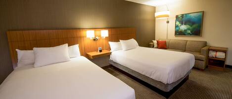 Quarto de hotel com duas camas, um sofá, uma mesa de cabeceira com abajur e um quadro na parede.