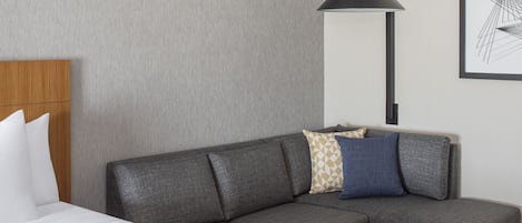 Een moderne hotelkamer met een grijze modulaire bank, een houten hoofdbord, een nachtkastje, een zwarte vloerlamp en een lijst met abstracte kunst aan de muur.