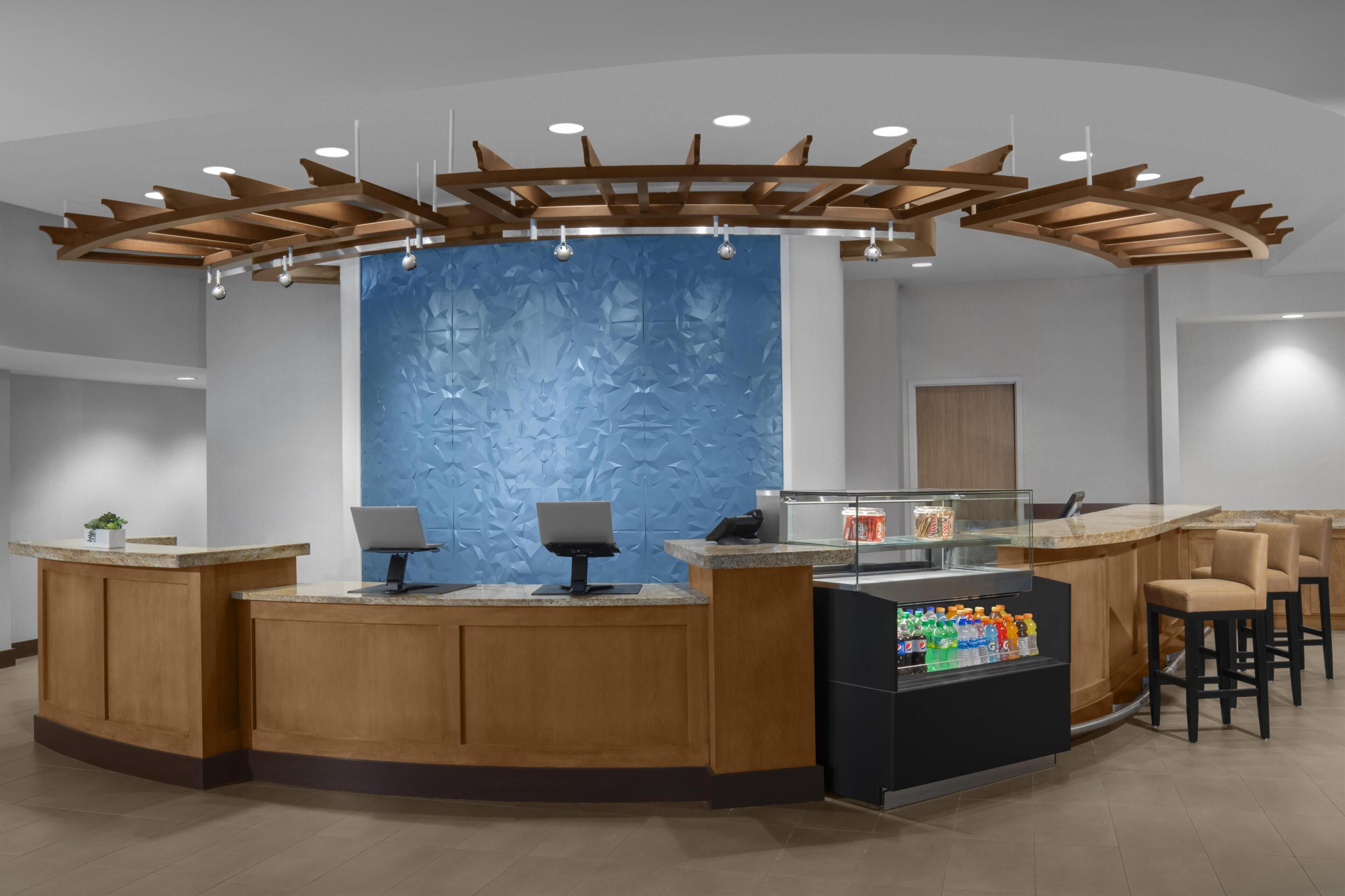 Un espace d’accueil avec un comptoir en bois, deux postes informatiques, un mur bleu à texture, et un plafond avec des poutres et des luminaires en bois.