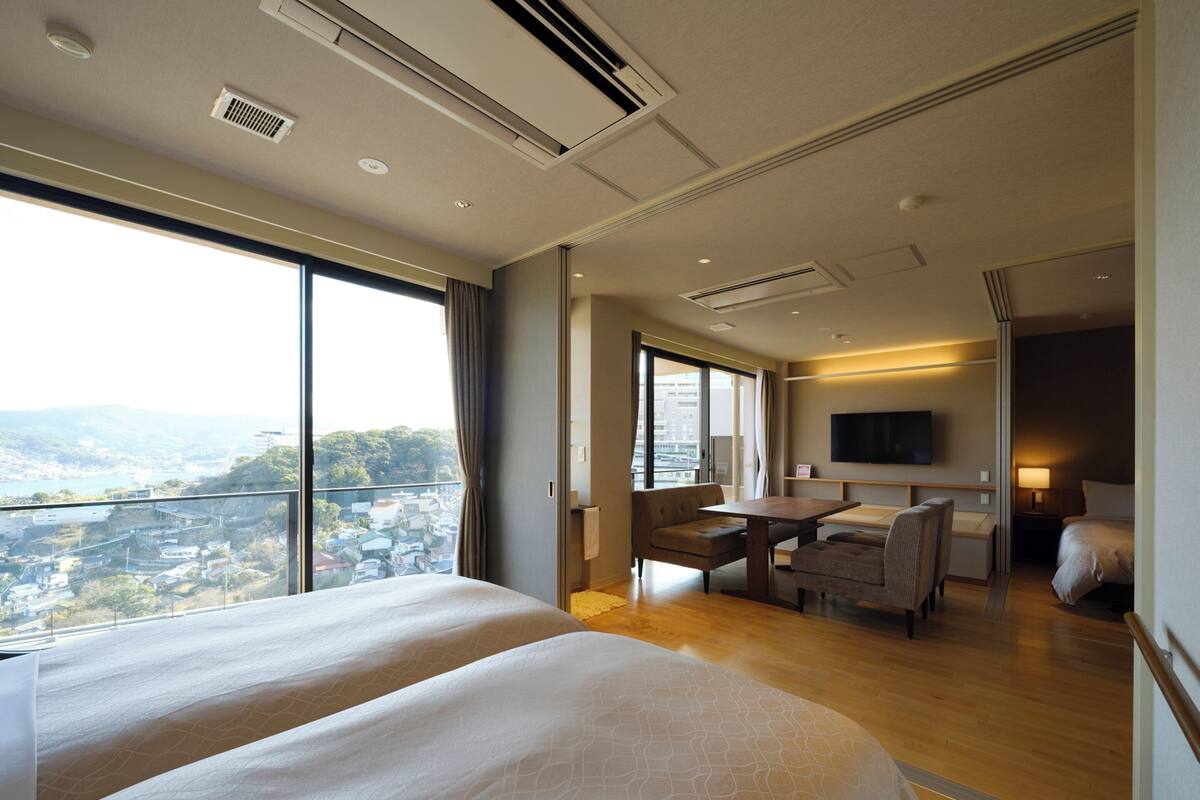 A hotel room with two beds, a desk, a chair, a TV, and a view of a cityscape.