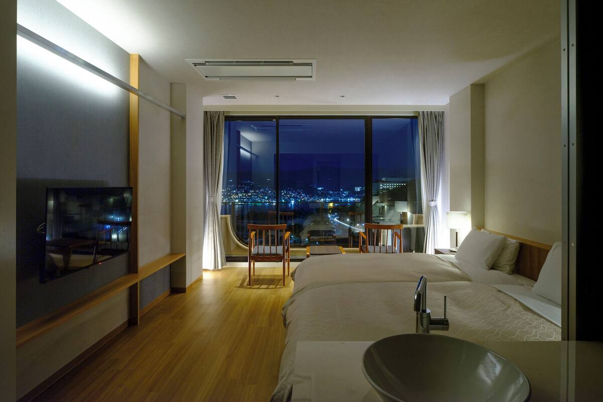 A modern hotel room with a large bed, a desk, and a view of a cityscape at night.