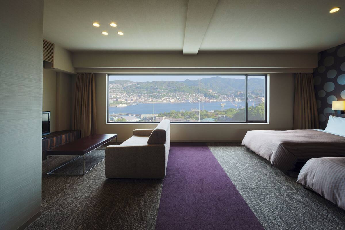 A hotel room with a large window offering a cityscape view, a bed, a desk, and a chair.