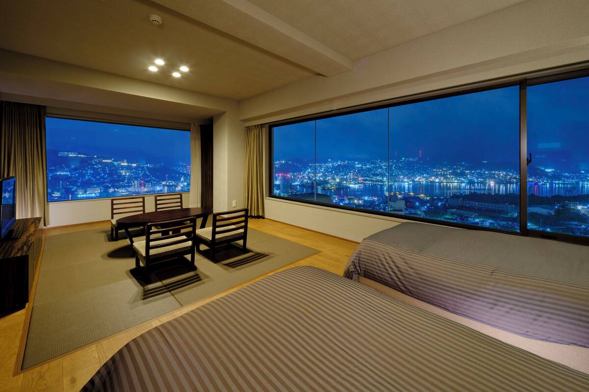A hotel room with a large window offering a city view at night.