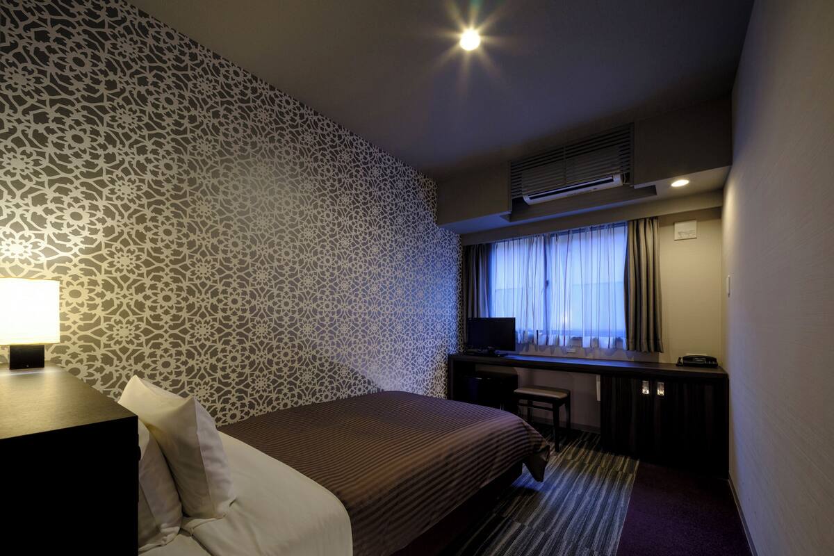 A hotel room with a patterned wall, a bed with pillows, a desk with a computer, and a window with curtains.