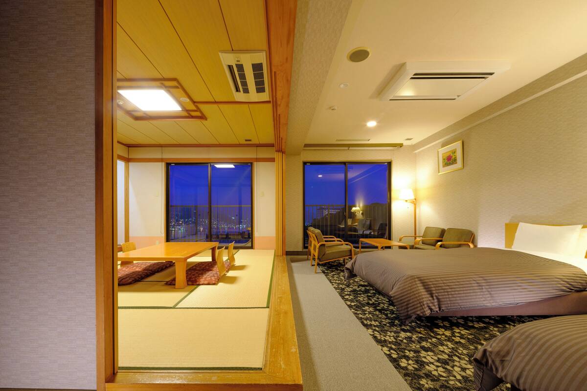 A hotel room with a traditional Japanese interior, featuring tatami flooring, a low wooden table, and a bed with a patterned bedspread.