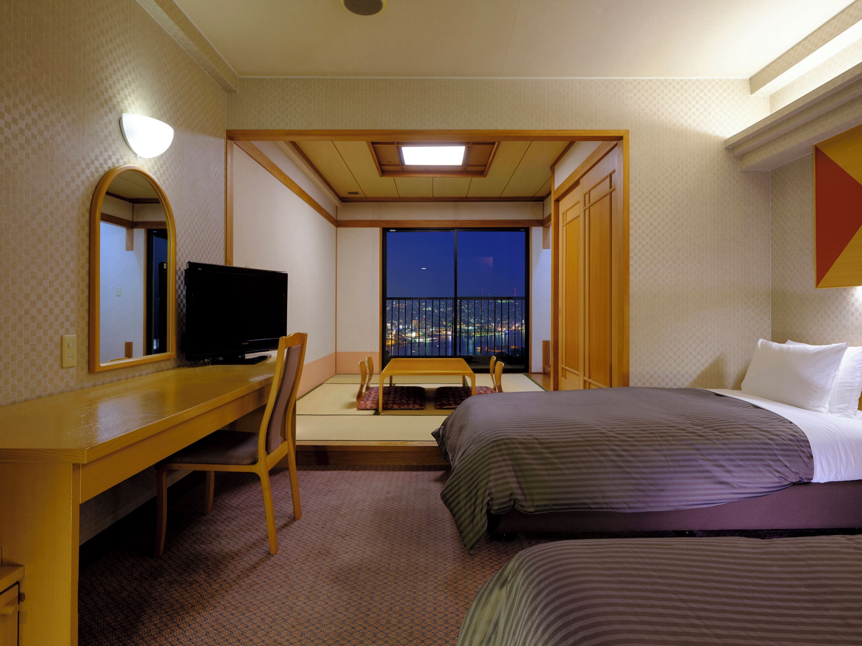 Een hotelkamer met een bureau, een televisie, een bed en uitzicht op de stad 's nachts.