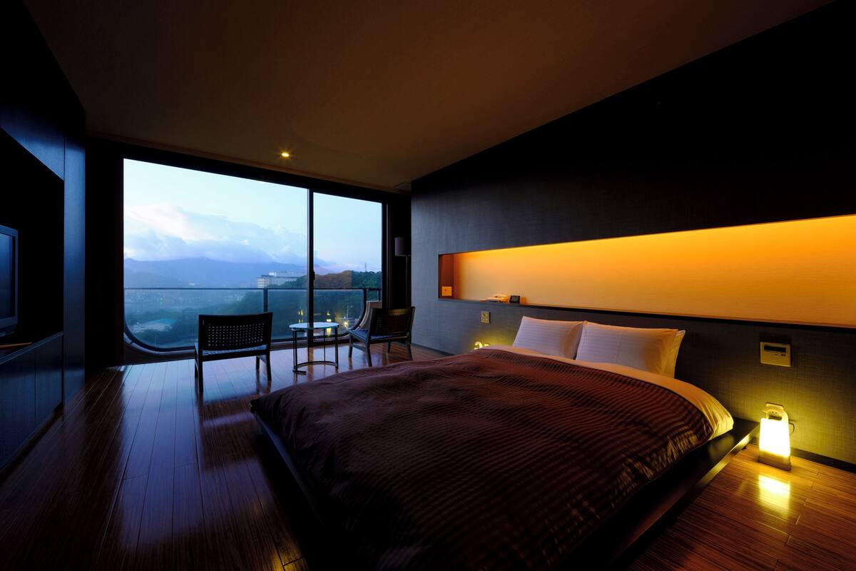 A modern hotel room with a large bed, a desk, and a chair by a large window offering a city view.