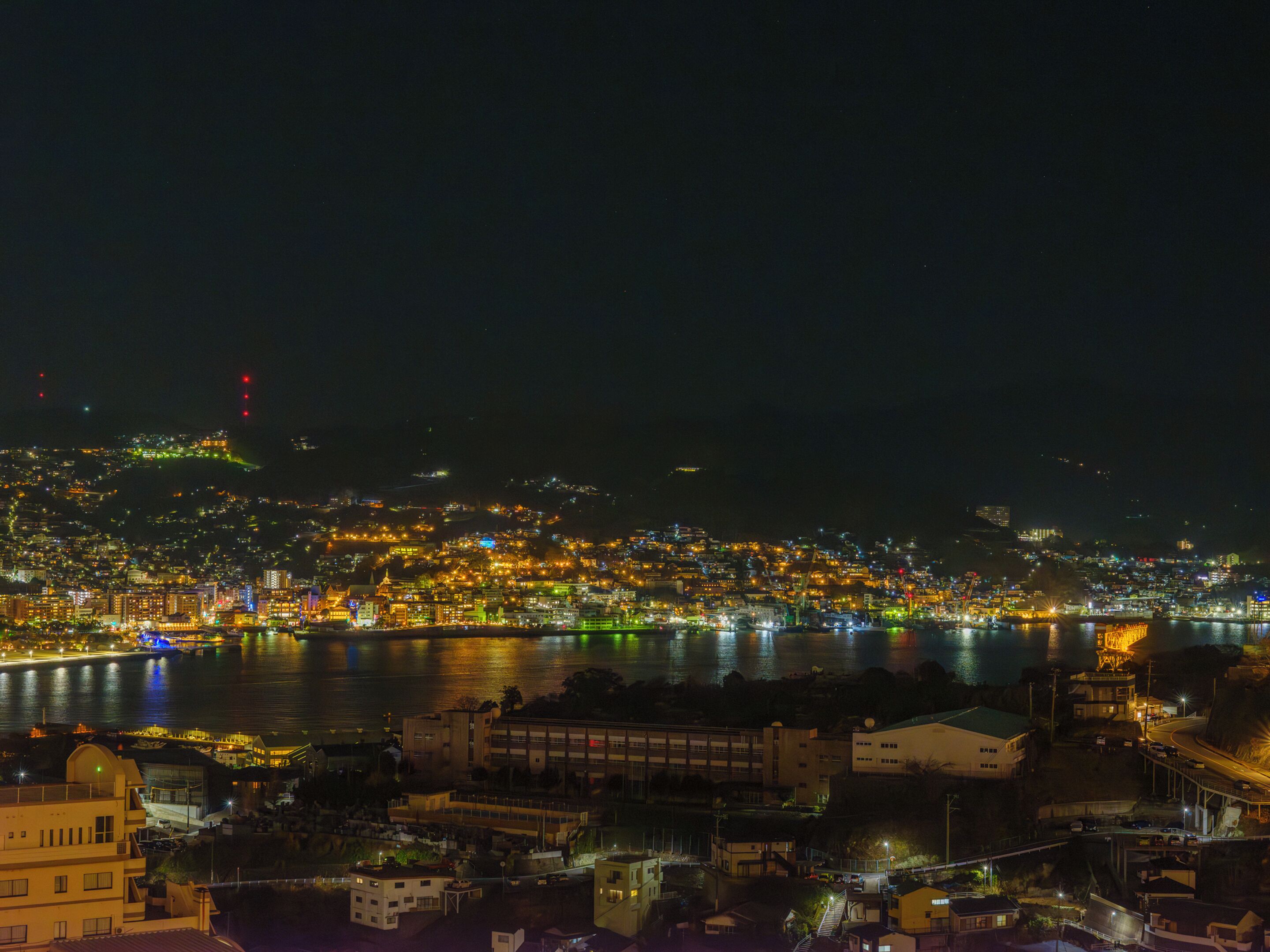 A nighttime cityscape with a river, buildings, and a bridge.