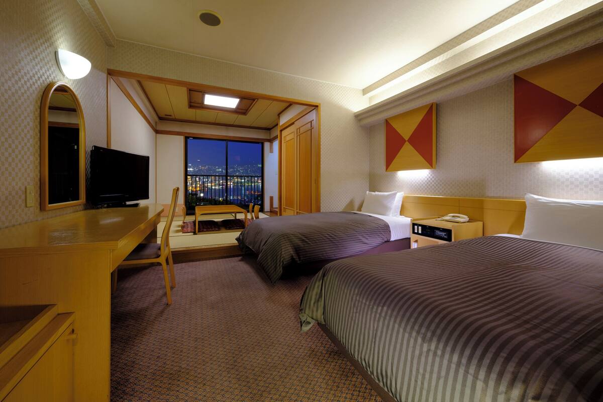 A hotel room with two beds, a desk, a TV, and a view of a cityscape at night.