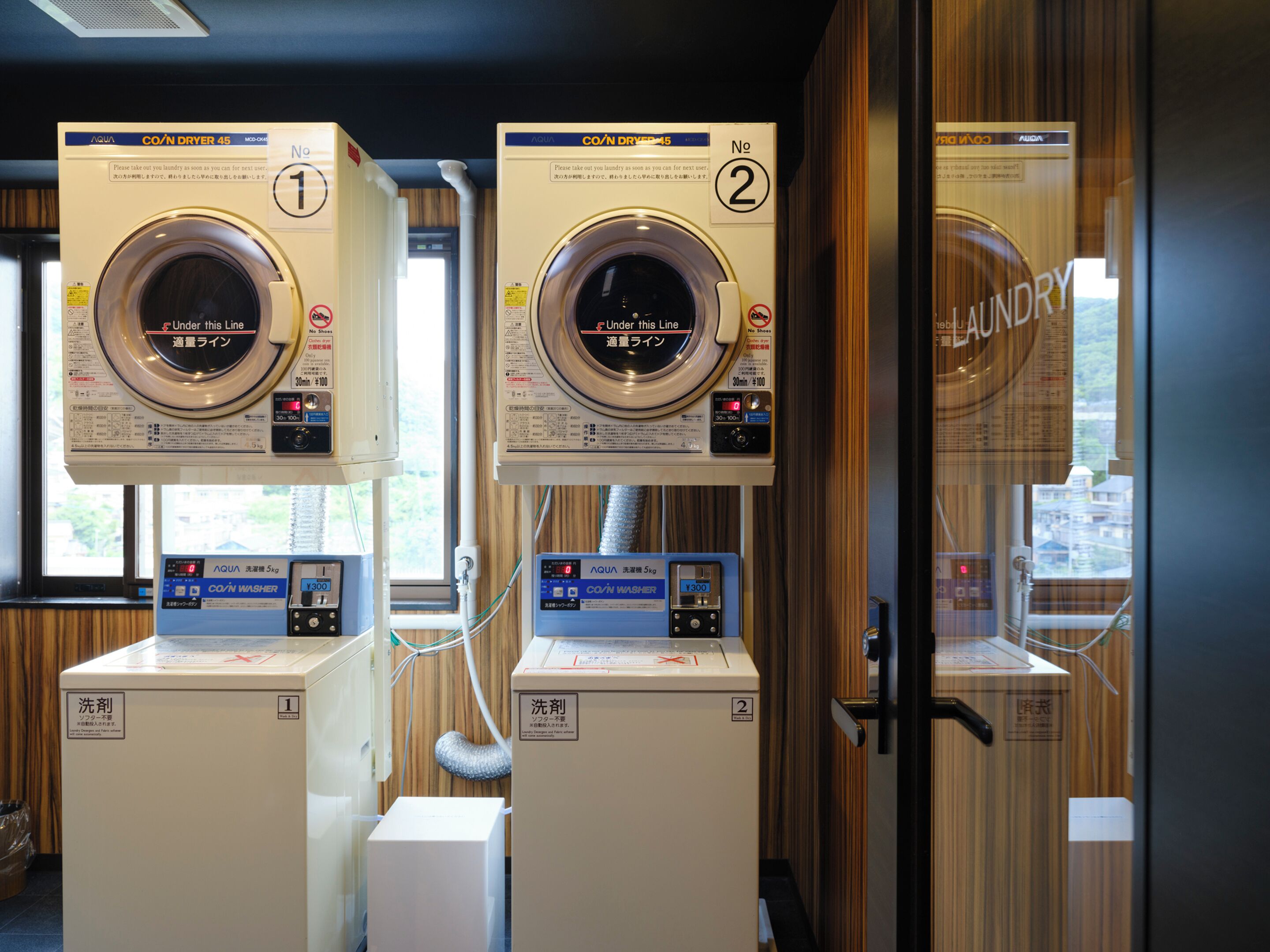 Two coin-operated laundry machines with digital displays, located in a room with wooden paneling and a window providing natural light.