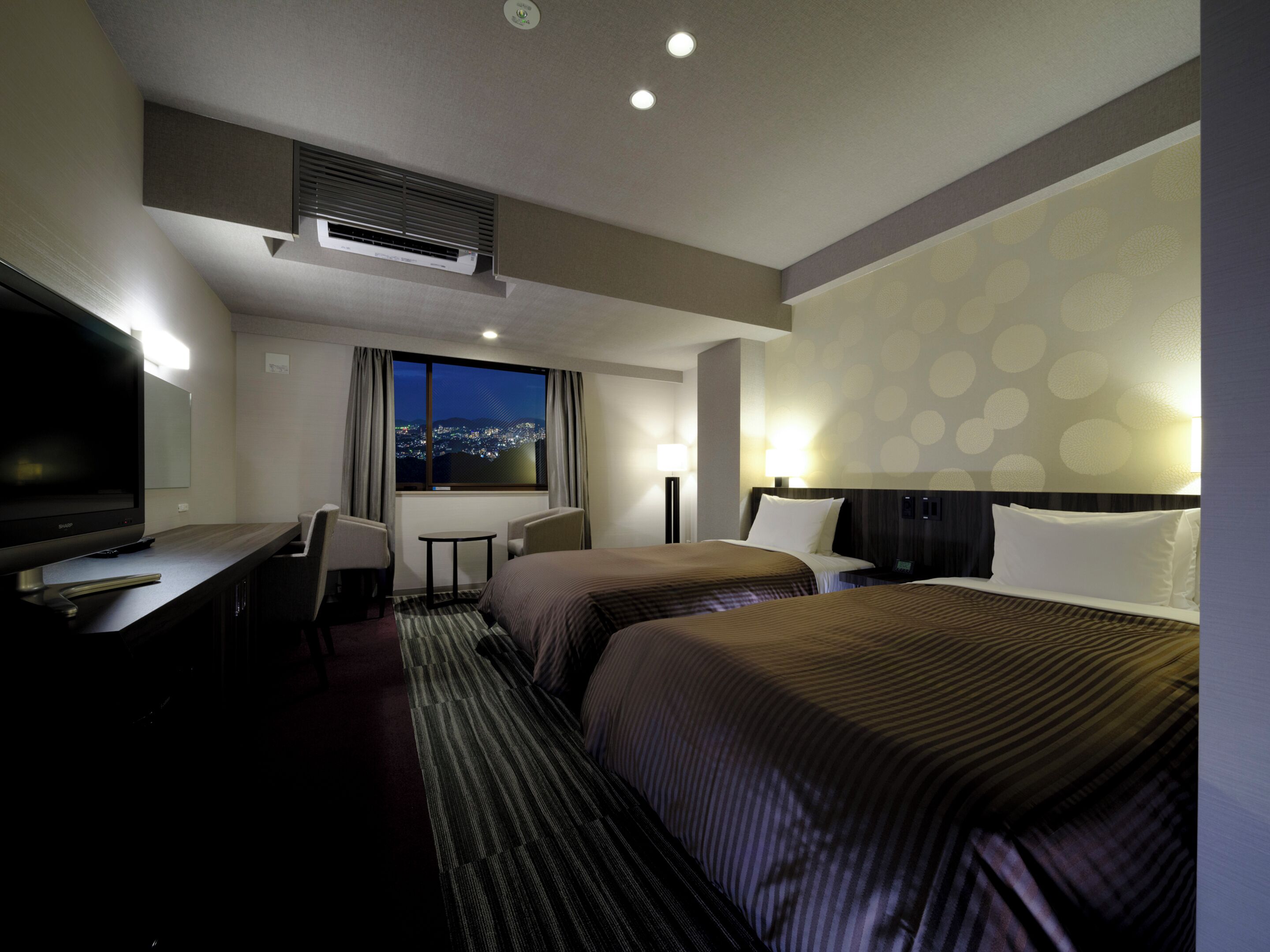 A hotel room with two beds, a desk, a TV, a window with a city view, and a patterned wall.