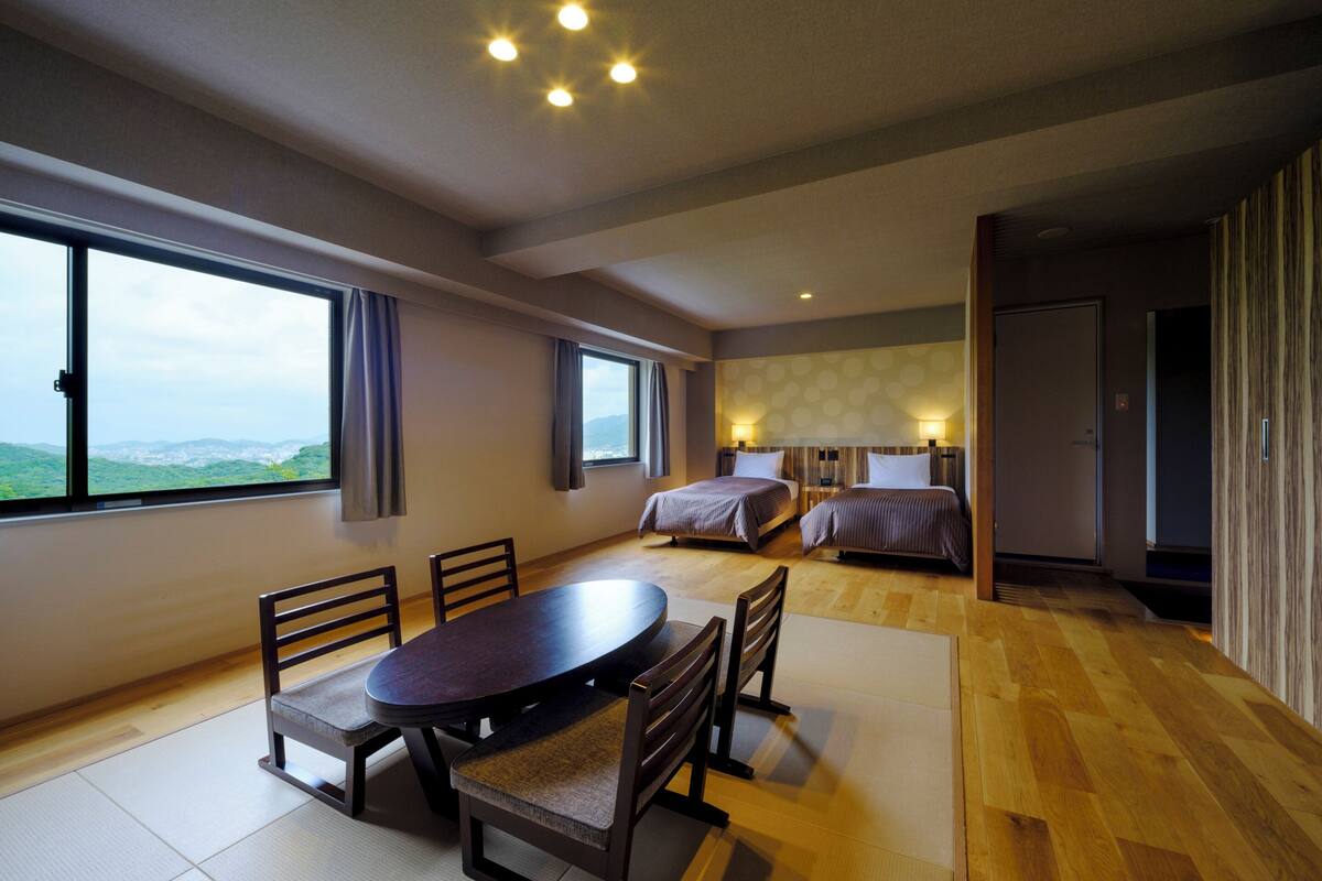 A hotel room with a large window, two beds, a dining table with chairs, and a view of the cityscape.