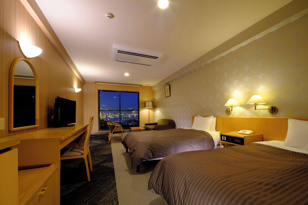 A hotel room with two beds, a desk with a TV, a chair, a nightstand with a phone, and a view of the city at night.