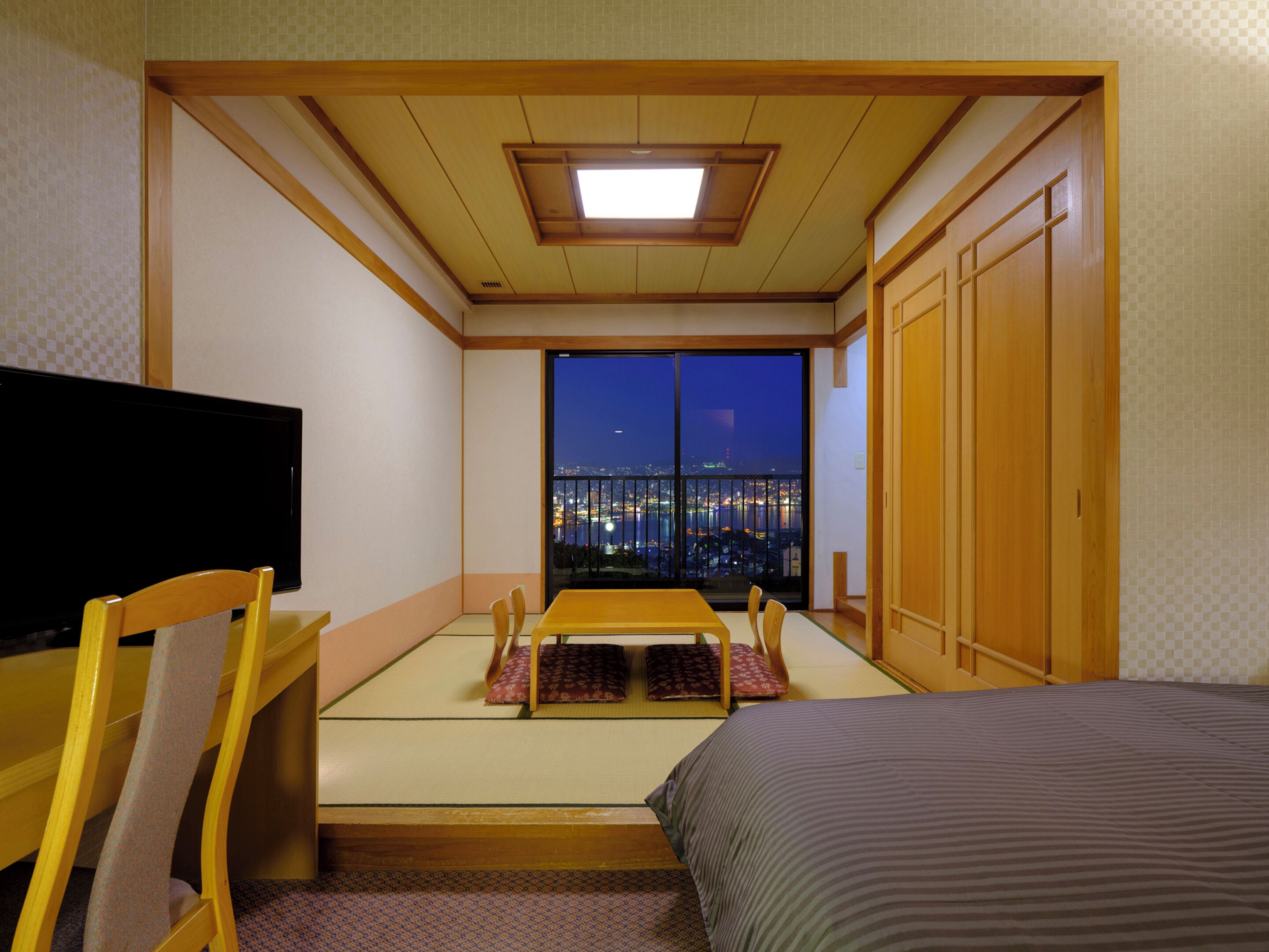Una habitación con cama, televisor, una mesa de comedor con sillas, vista a la ciudad de noche y una puerta corrediza.