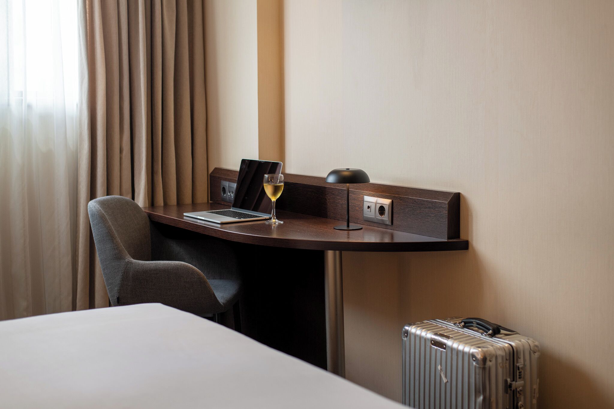 Een hotelkamer met een bureau, een laptop, een glas wijn, een koffer en een stoel.
