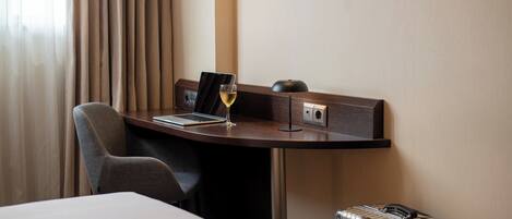 A hotel room with a desk, a laptop, a glass of wine, a suitcase, and a chair.