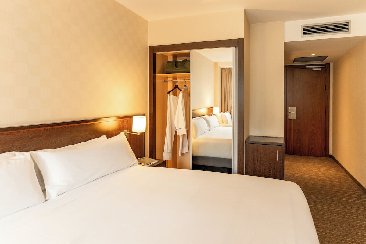 A hotel room with a large bed, a bedside table with a lamp, a wardrobe, and a door leading to another room.