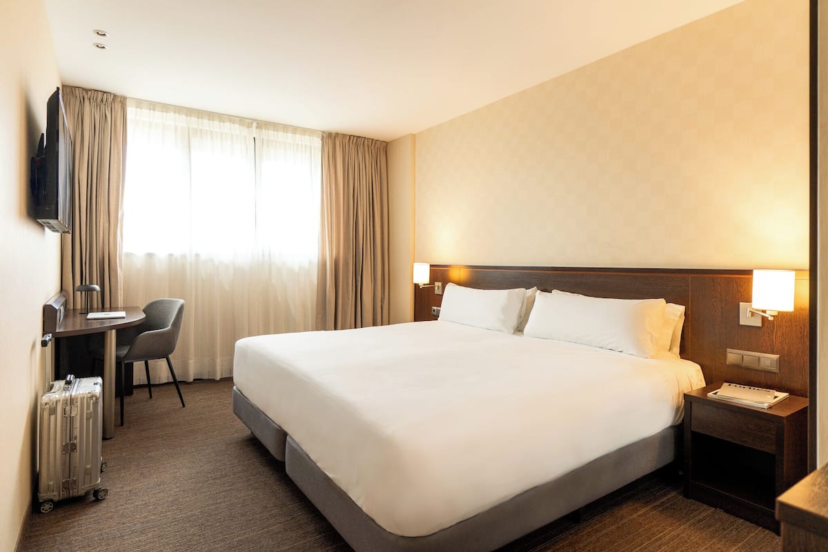 A hotel room with a large bed, a desk with a chair, a television, and a suitcase.