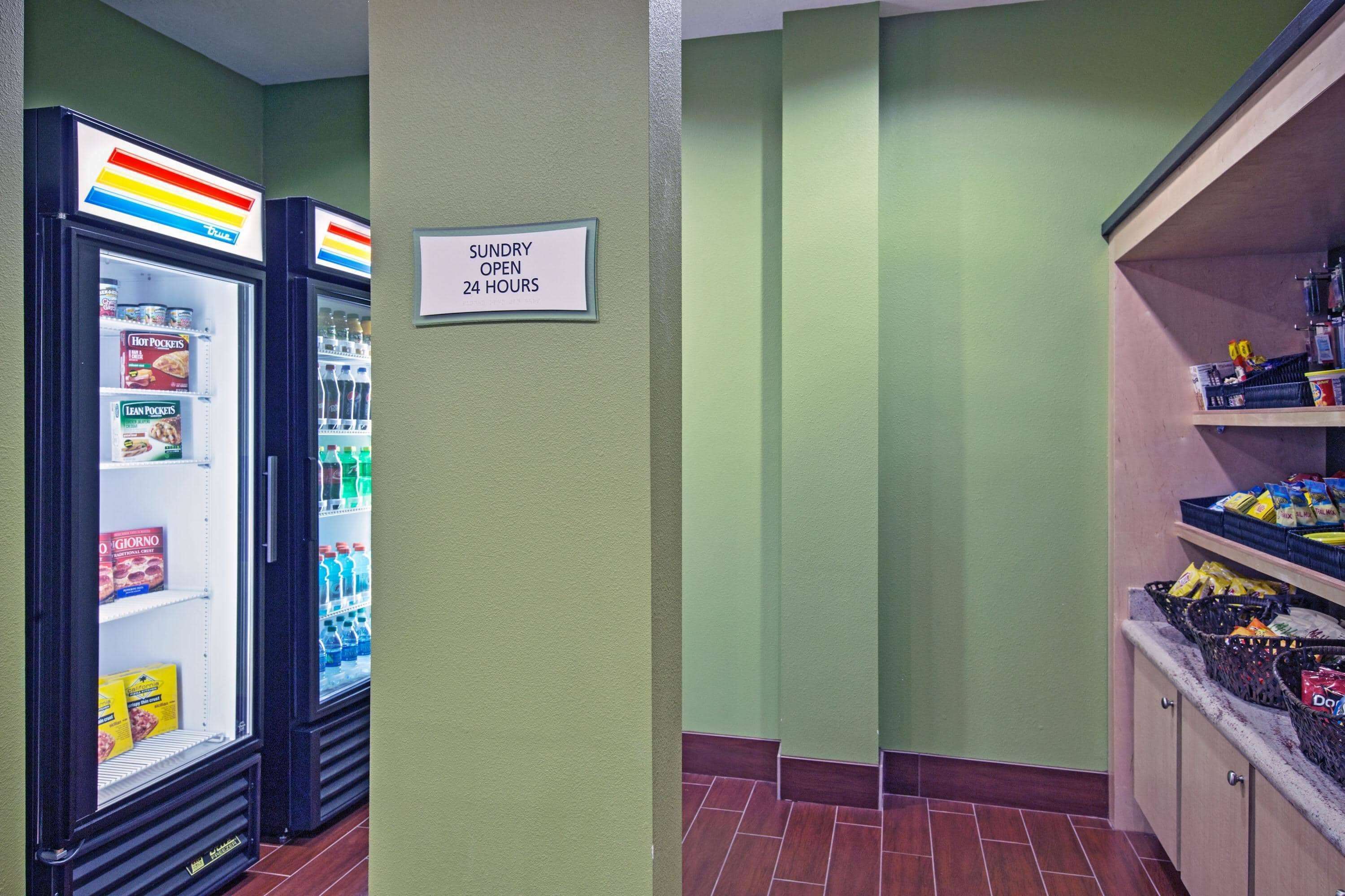 A kitchenette area with two refrigerators stocked with food items and a sign indicating the sundry is open 24 hours.