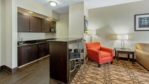 A compact hotel room with a kitchenette, a small dining area, a patterned carpet, and a single orange armchair. - La Quinta Inn & Suites by Wyndham Leesville Ft. Polk (Leesville)