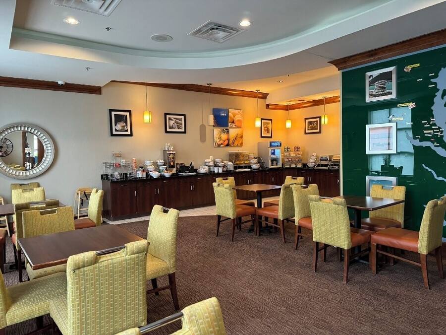 Un comedor de hotel con mesas redondas, sillas amarillas, un mostrador de buffet y una pared con cuadros enmarcados.