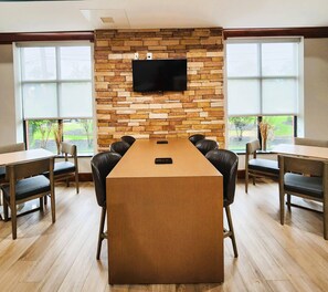 A conference room with a long table, six chairs, a flat-screen TV mounted on a stone wall, and large windows with a view of greenery.