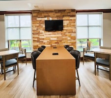 A conference room with a long table, six chairs, a flat-screen TV mounted on a stone wall, and large windows with a view of greenery.