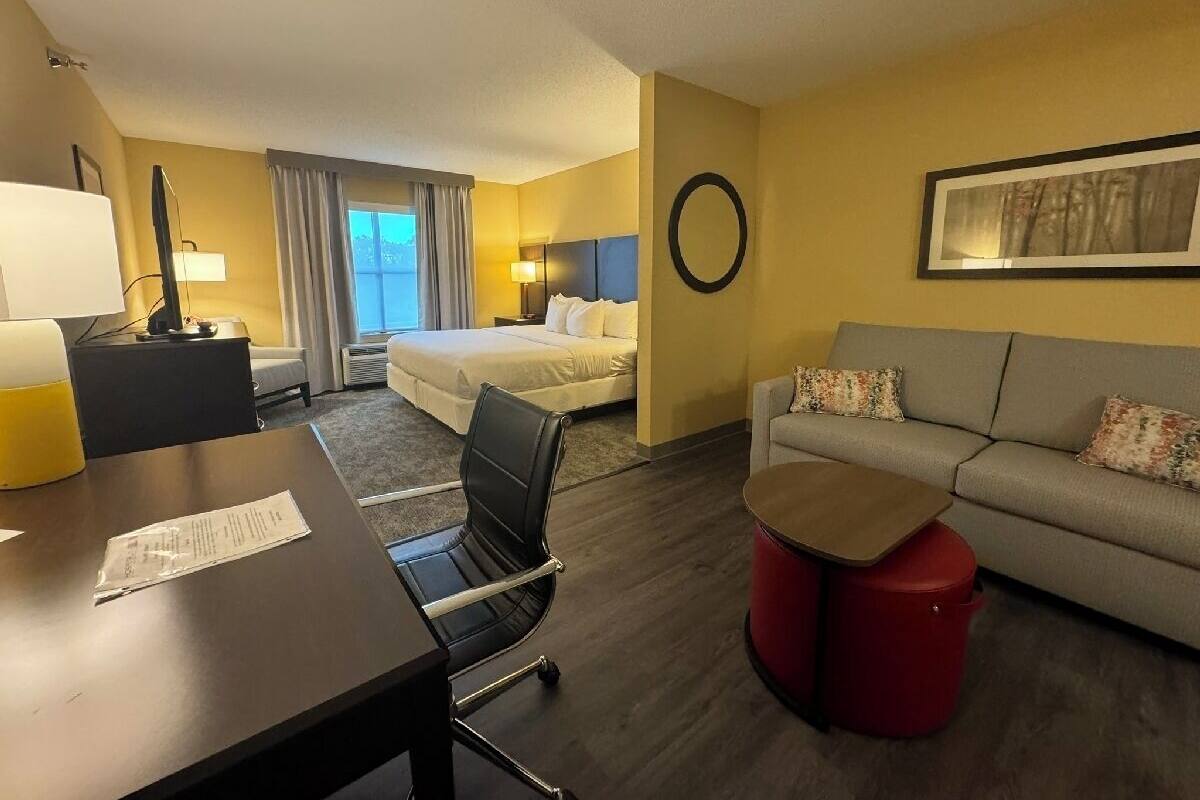 A hotel room with a bed, a sofa, a desk with a chair, a lamp, and a round table.