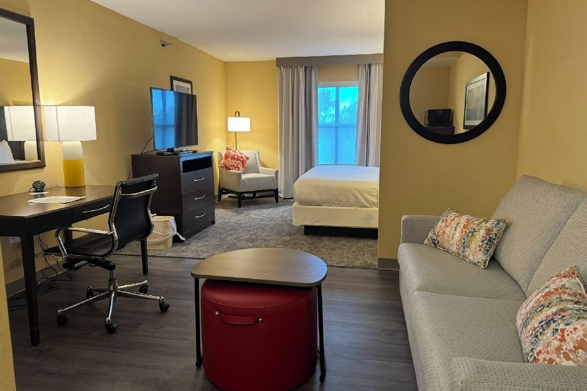 A hotel room with a desk, office chair, a red ottoman, a sofa, a bed, a TV, and a mirror.