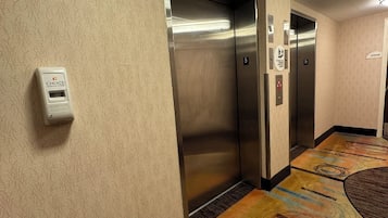 A hotel corridor with two elevators, a wall-mounted intercom, and a carpeted floor.