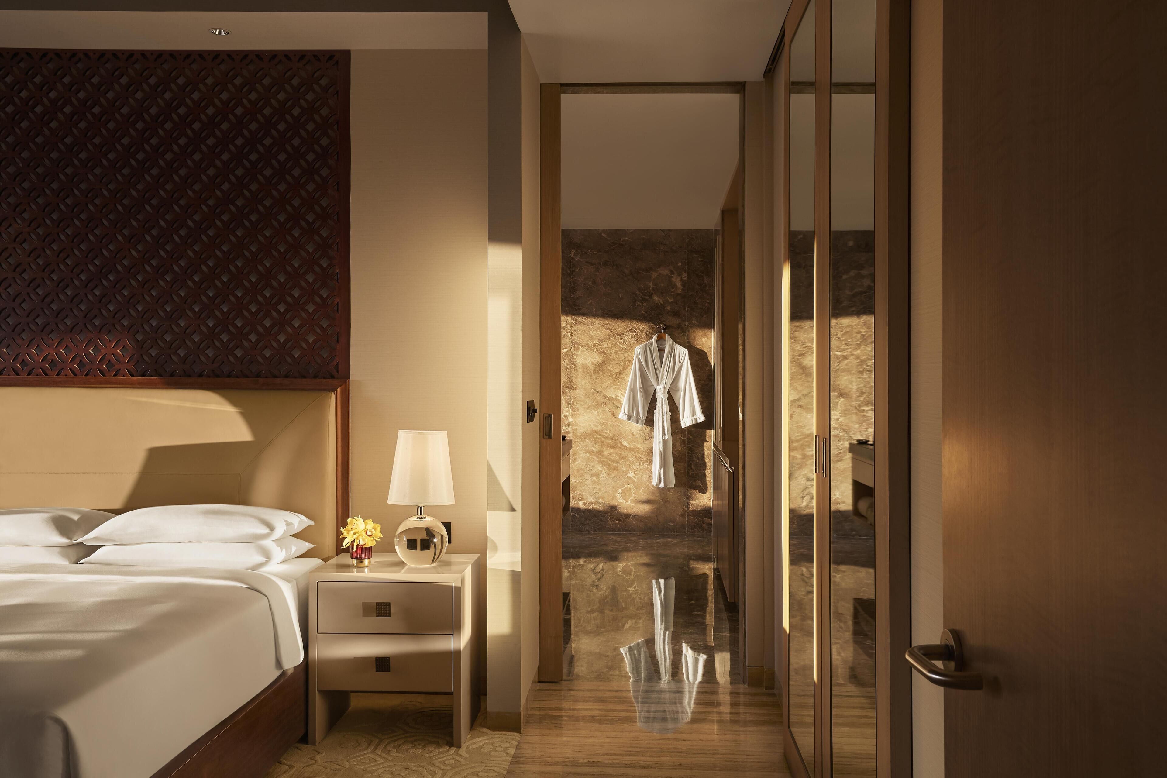 A hotel room with a bed, bedside table, lamp, and a view of a hallway with a robe hanging on a hook.