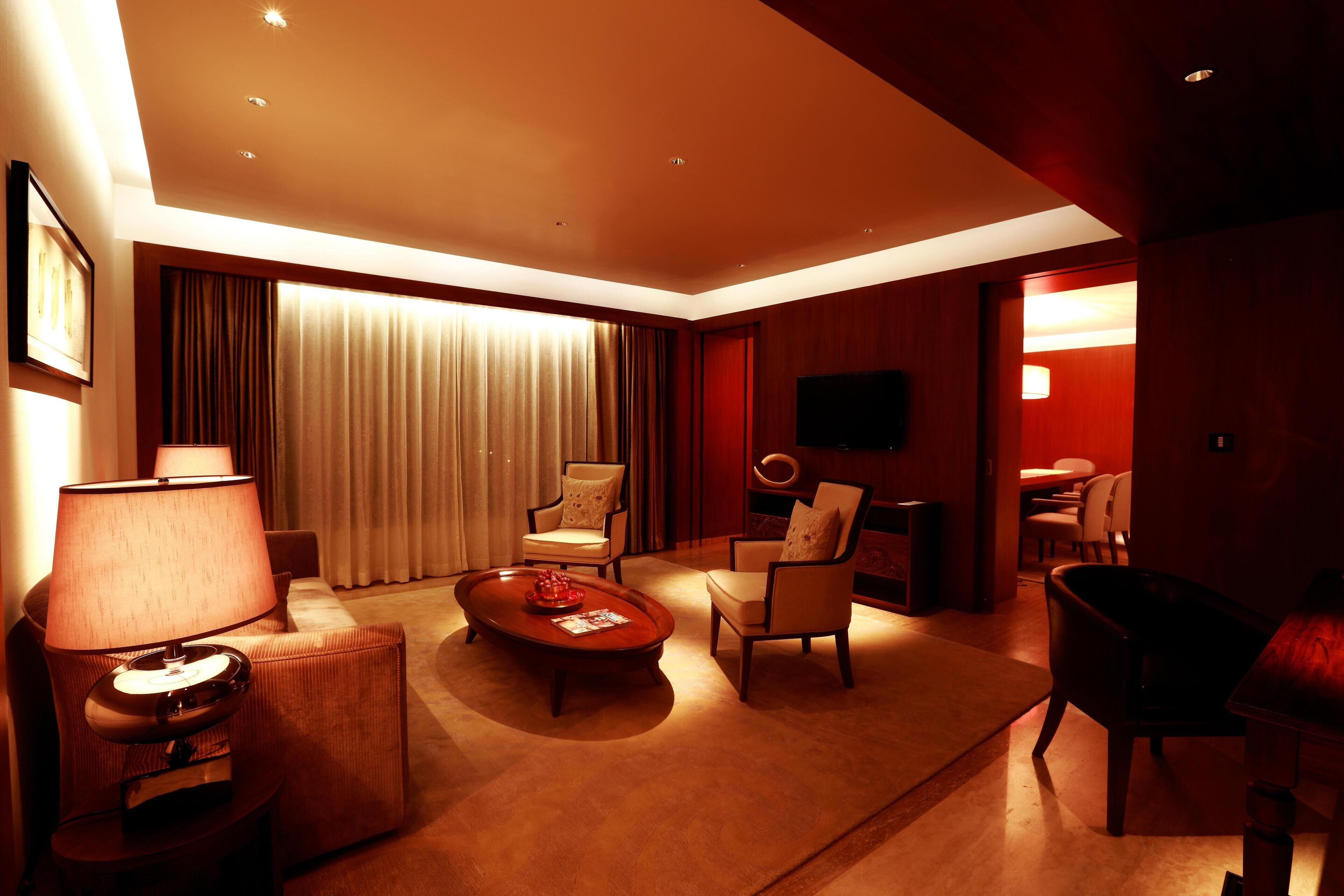 Habitación de hotel con sofá, sillón y mesa de centro. Hay un televisor montado en la pared y una ventana con cortinas.