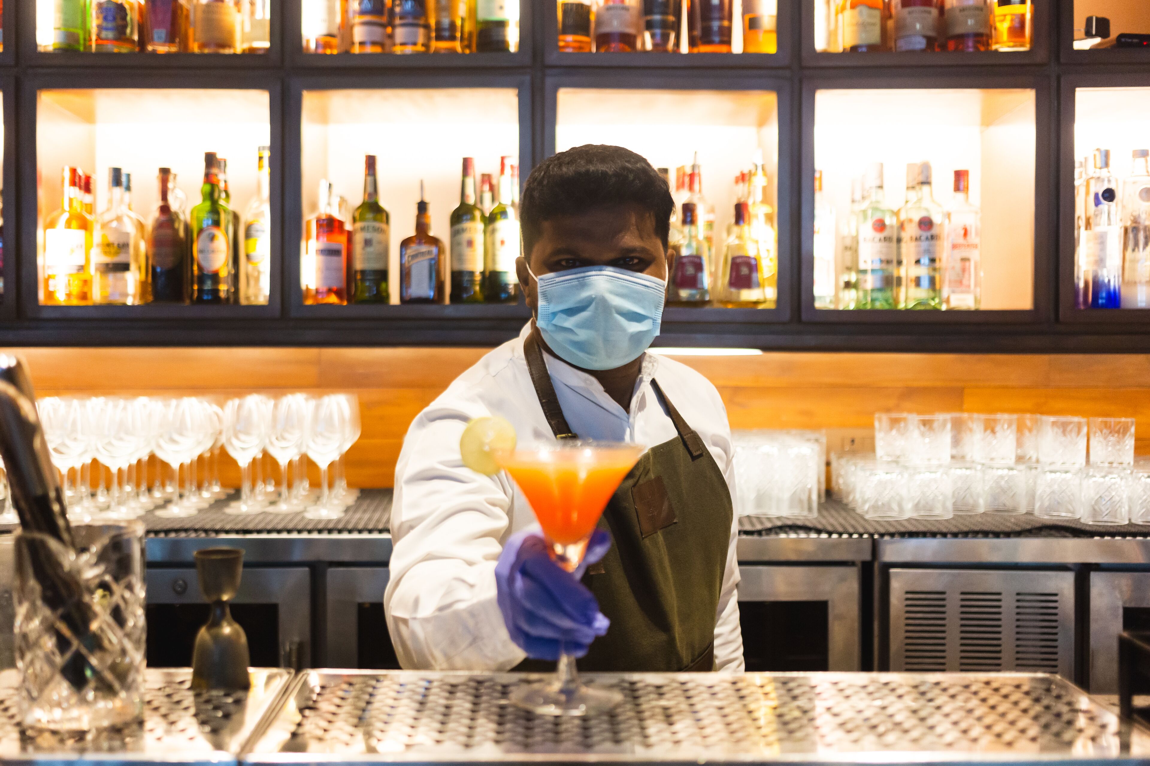 Un barman sosteniendo un cóctel con una rodaja de lima, detrás de una barra con estantes llenos de botellas de licor y vasos.