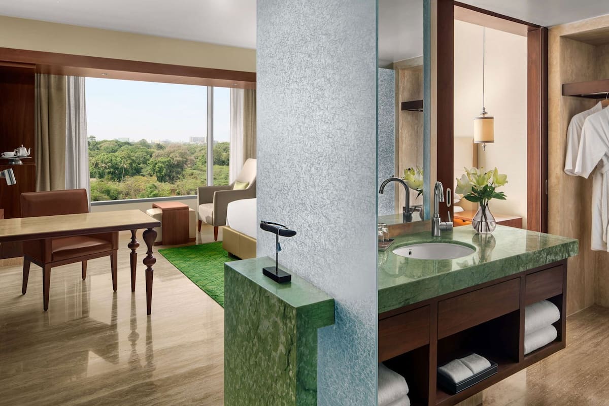 Hotelkamer met een eethoek, een groot raam met uitzicht op de stad, een badkamer met een wastafel en spiegel, en een groen tapijt.
