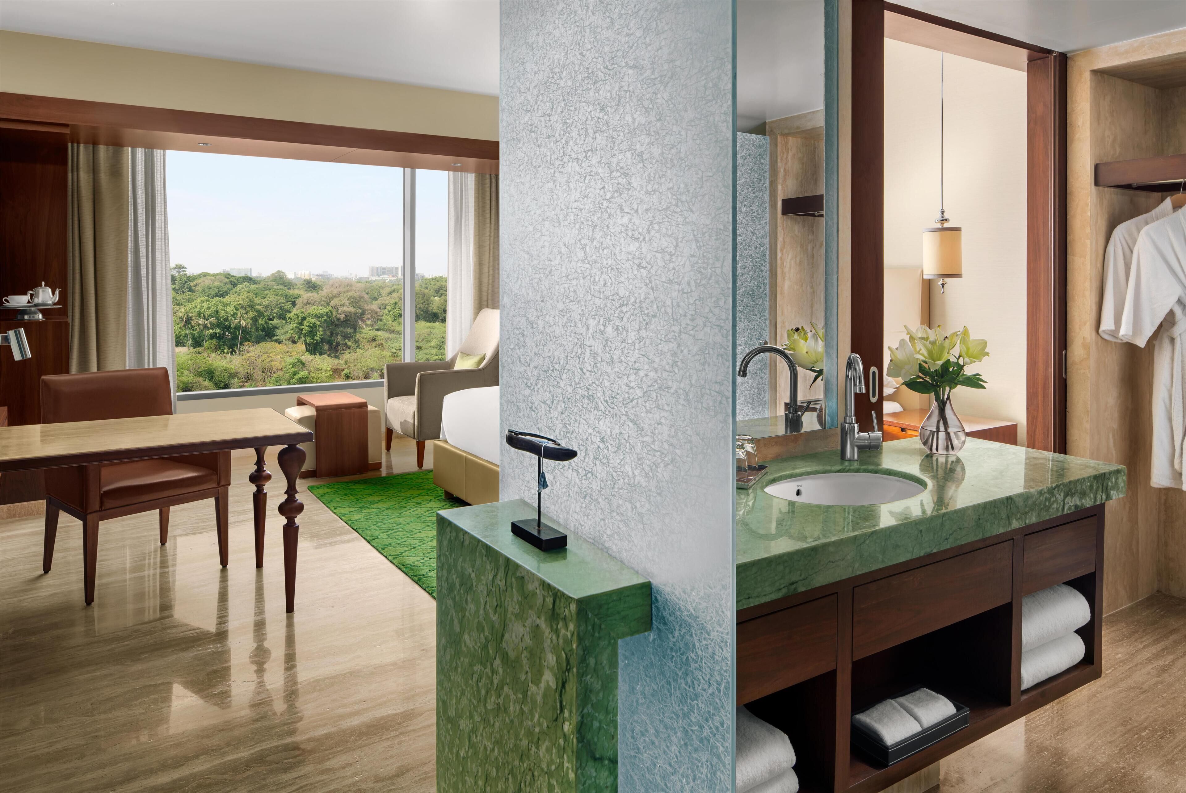 A hotel room with a large window, a green marble bathroom vanity, a wooden dining table, and a view of greenery outside.