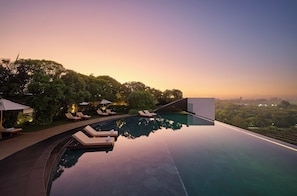 Ein Infinity-Pool mit Liegestühlen und Sonnenschirmen, umgeben von üppiger Vegetation und einer Stadtkulisse im Hintergrund.
