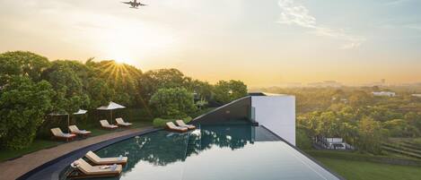 A modern infinity pool with lounge chairs and umbrellas, surrounded by lush greenery and a cityscape in the background.