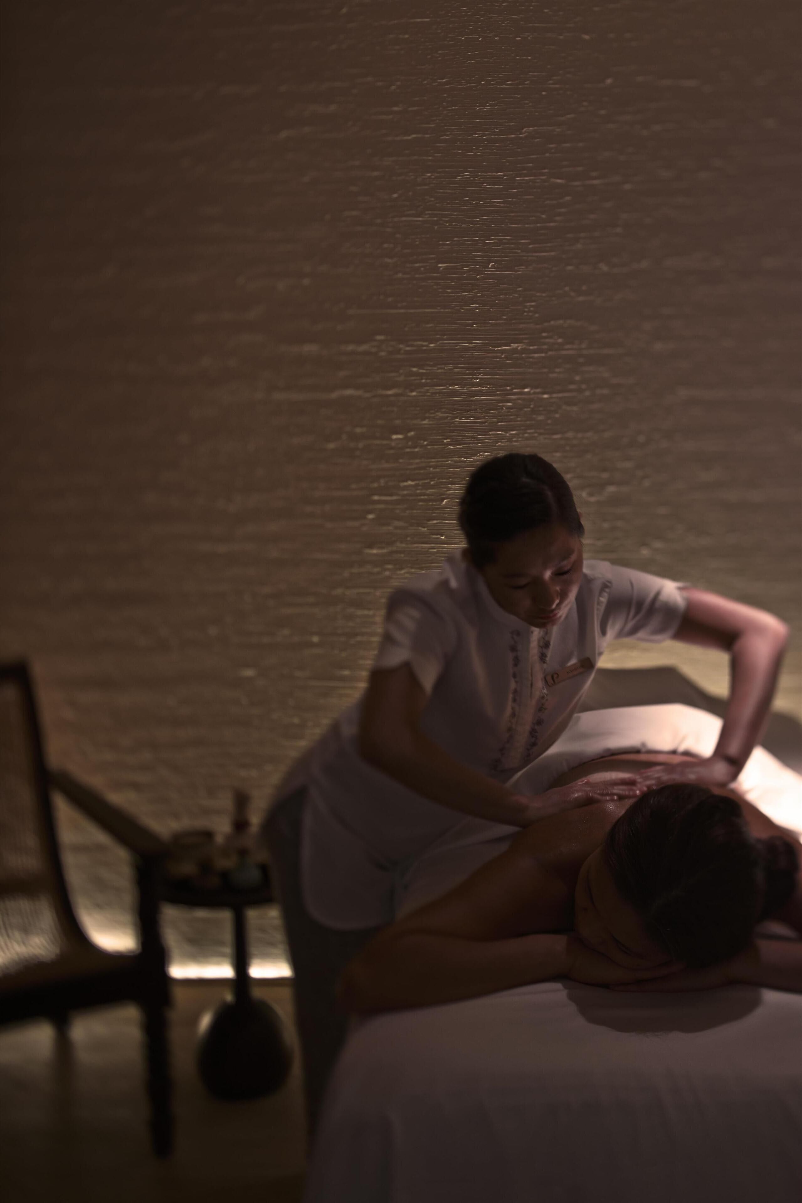 A person receiving a massage in a dimly lit room with a bed and a small table with a bottle.